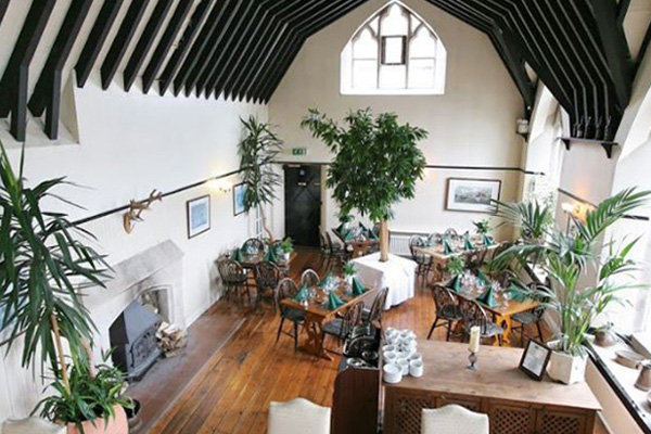 spacious dining area with high ceiling and large windows featuring wooden beams tables set for two with greenery and a fireplace